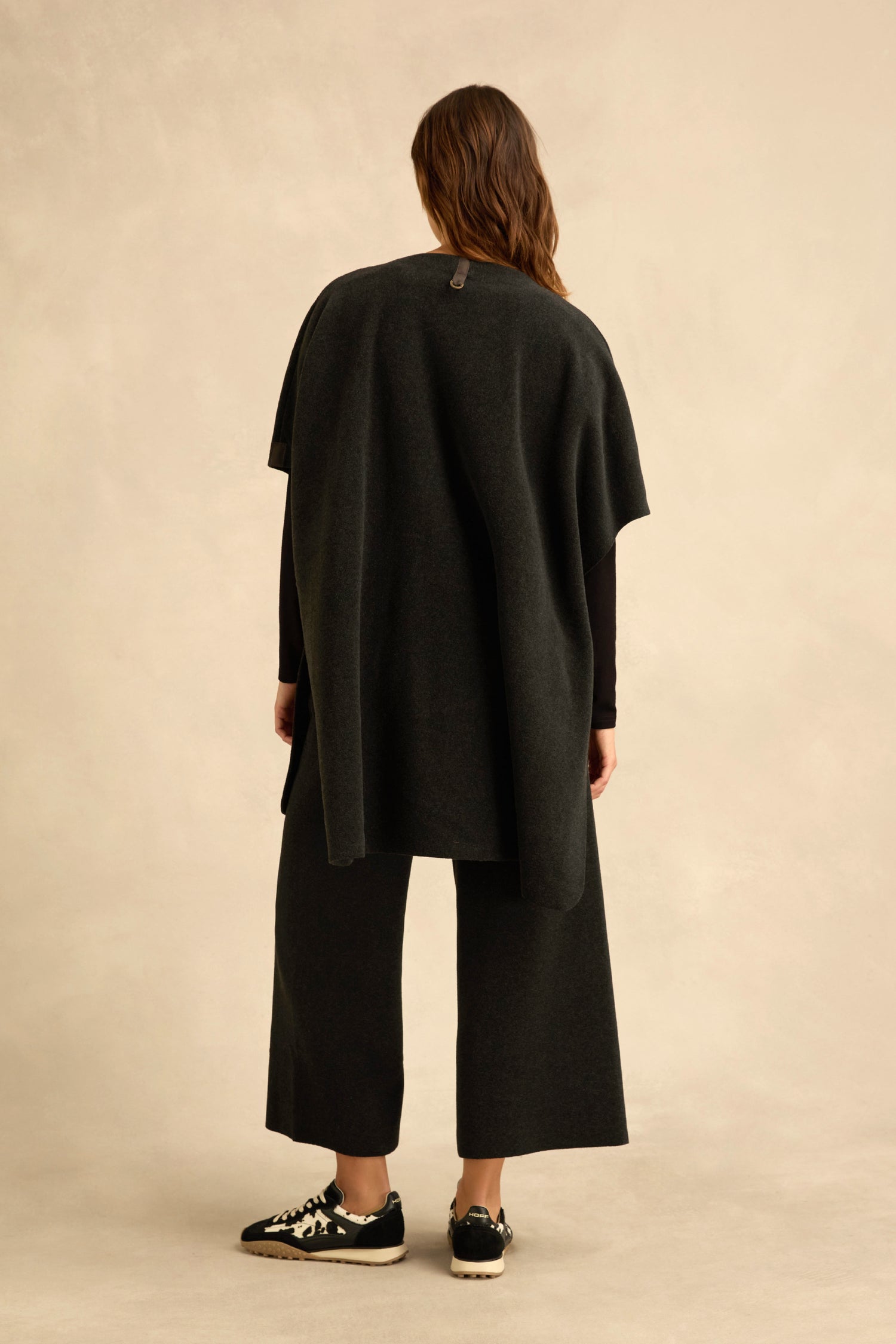 A person stands with their back to the camera, wearing the Short Sleeve Fleece Gilet styled with loose dark clothing and kimono-style sleeves, paired with black sneakers, against a neutral background.