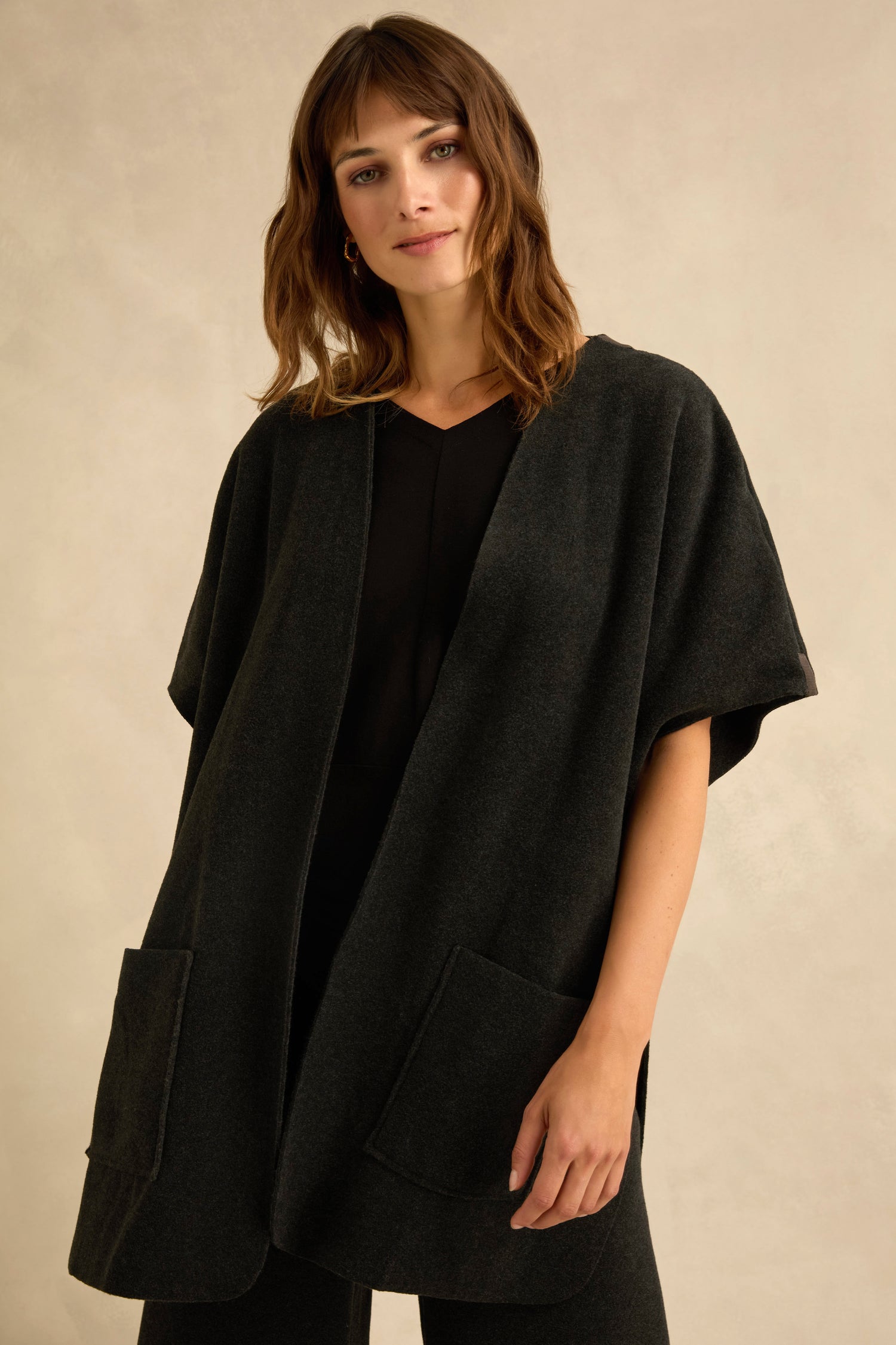 A woman wearing a dark, short sleeve fleece gilet with a loose fit and large front pockets over a black top, standing against a neutral background.