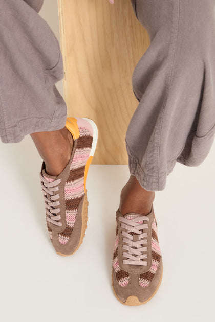 Person wearing taupe wide-leg pants and Bridge Woven Brown Crochet Trainers with a cushioned sole and pink, brown, and yellow accents, standing near a wooden block.