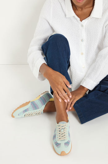 Wearing a white textured shirt, blue pants, and Bridge Woven Light Blue Crochet Trainers with a cushioned sole, a person sits on the floor with one knee up and hands resting on their ankle.