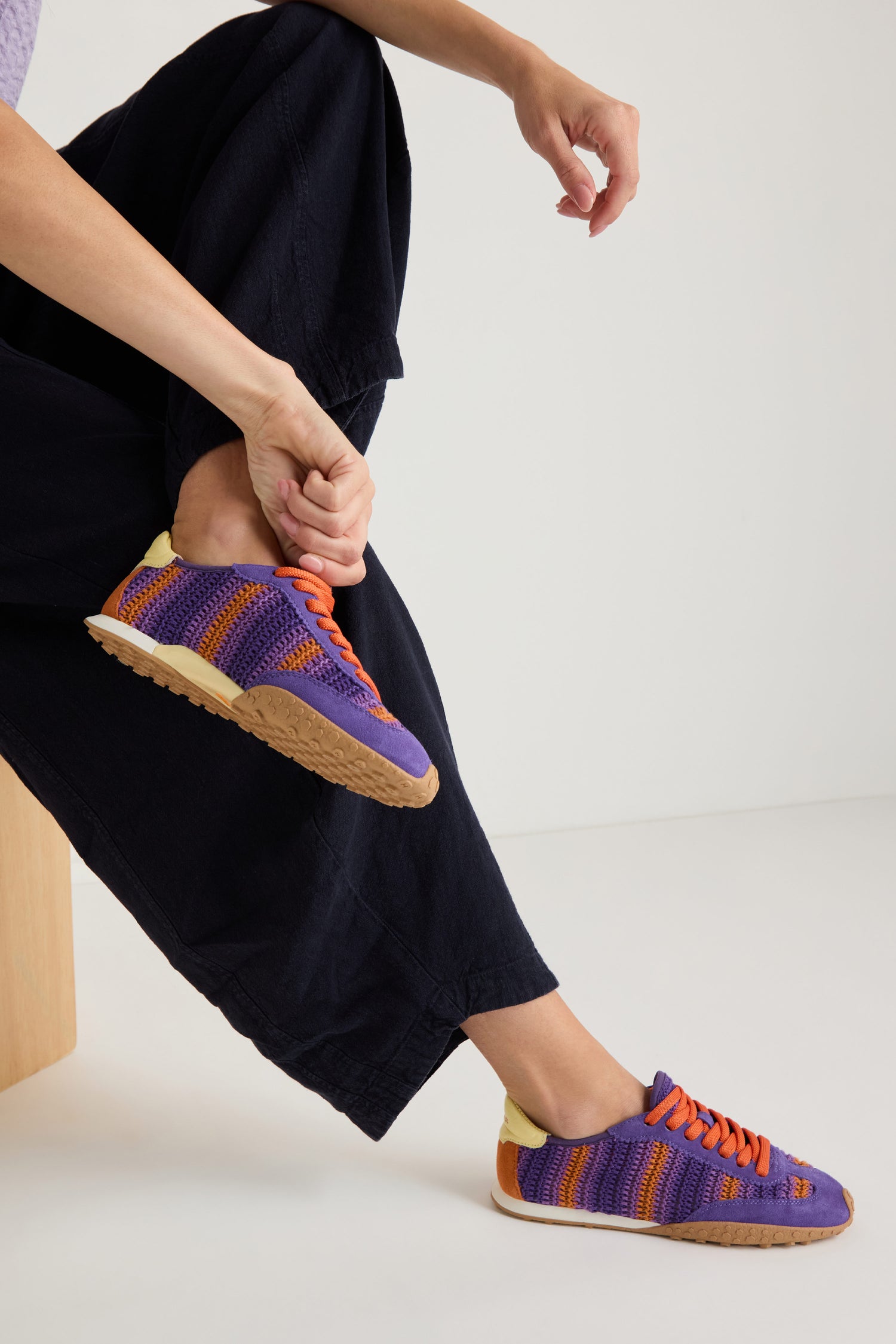 A person sits on a wooden stool in black pants, lacing up Bridge Woven Purple Crochet Trainers featuring colorful purple, orange, and yellow patterns and a cushioned sole against a plain background.