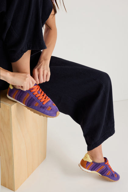 Seated on a wooden block, a person in black pants ties the orange laces of their Bridge Woven Purple Crochet Trainers, which have purple and orange panels and a cushioned sole for comfort.