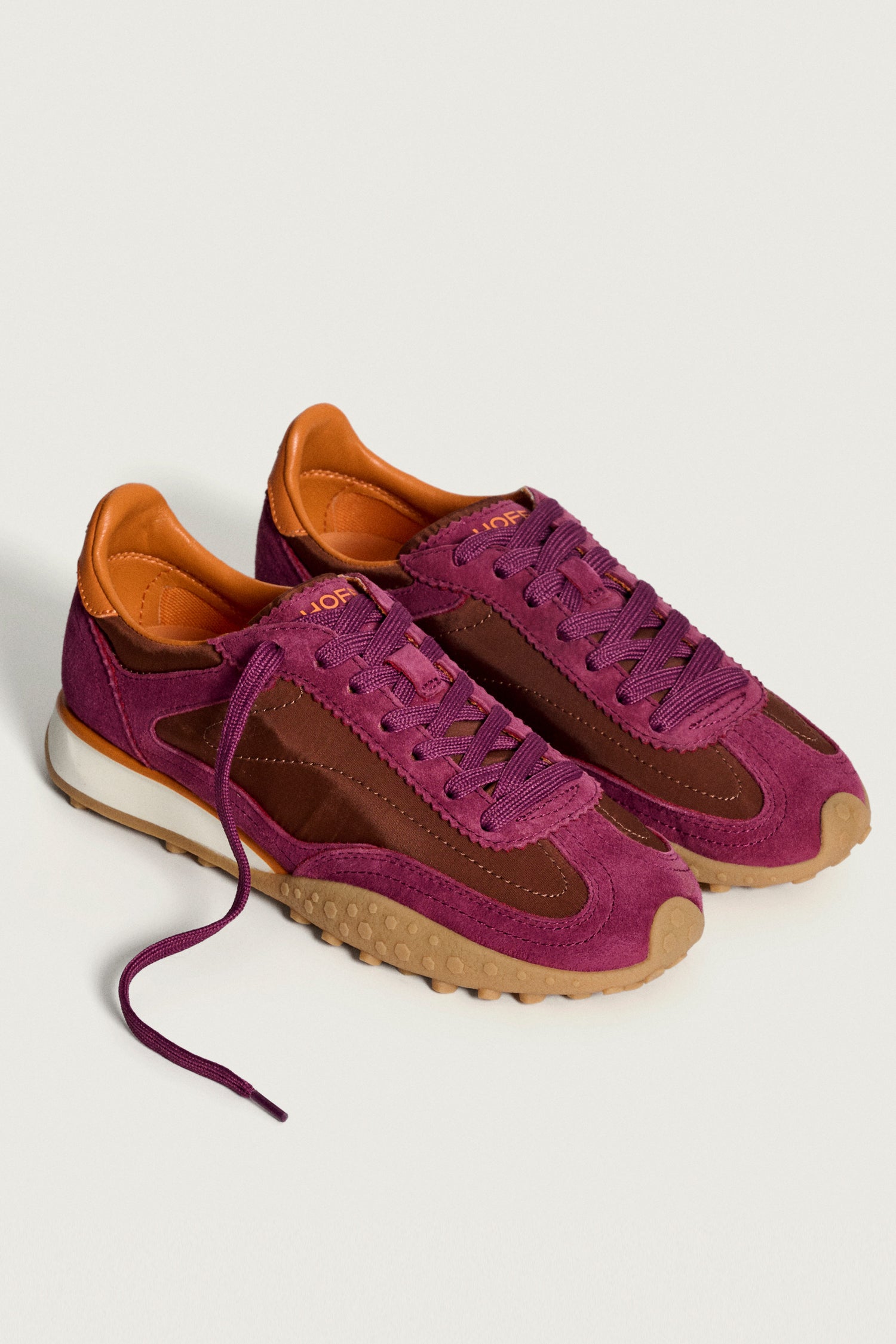 The Burgundy Bridge Trainers feature purple and brown suede with tan accents, orange lining, and gum rubber soles, shown on a plain white background. These modern essentials are perfect for comfortable everyday wear.