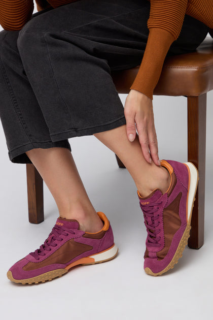 A person sits on a chair in black pants, an orange-brown sweater, and Burgundy Bridge Trainers—purple-orange sneakers—while touching their ankle.