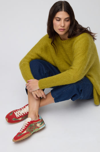 A woman in a yellow sweater and blue pants sits cross-legged on the floor, looking down, wearing Red William Morris Bridge Trainers.