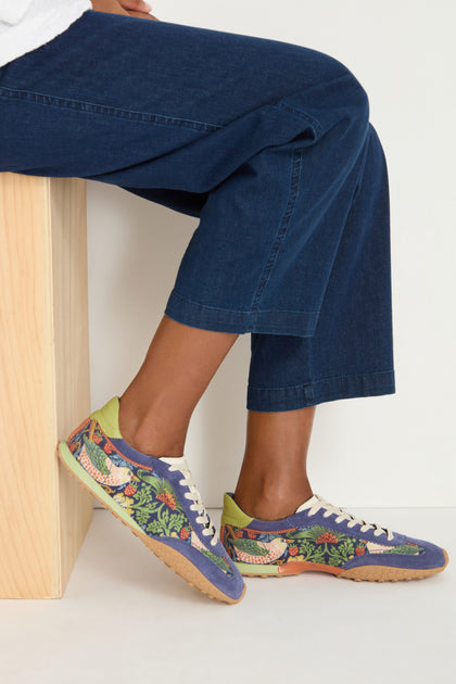 A person sits on a wooden bench, wearing dark blue jeans and vibrant William Morris Bridge Trainers featuring the Strawberry Thief print, white laces, and a cork-texture sole.