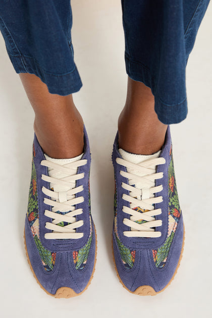 A person wearing William Morris Bridge Trainers with blue patterns, white laces, and tan soles stands on a light surface, dressed in blue pants.