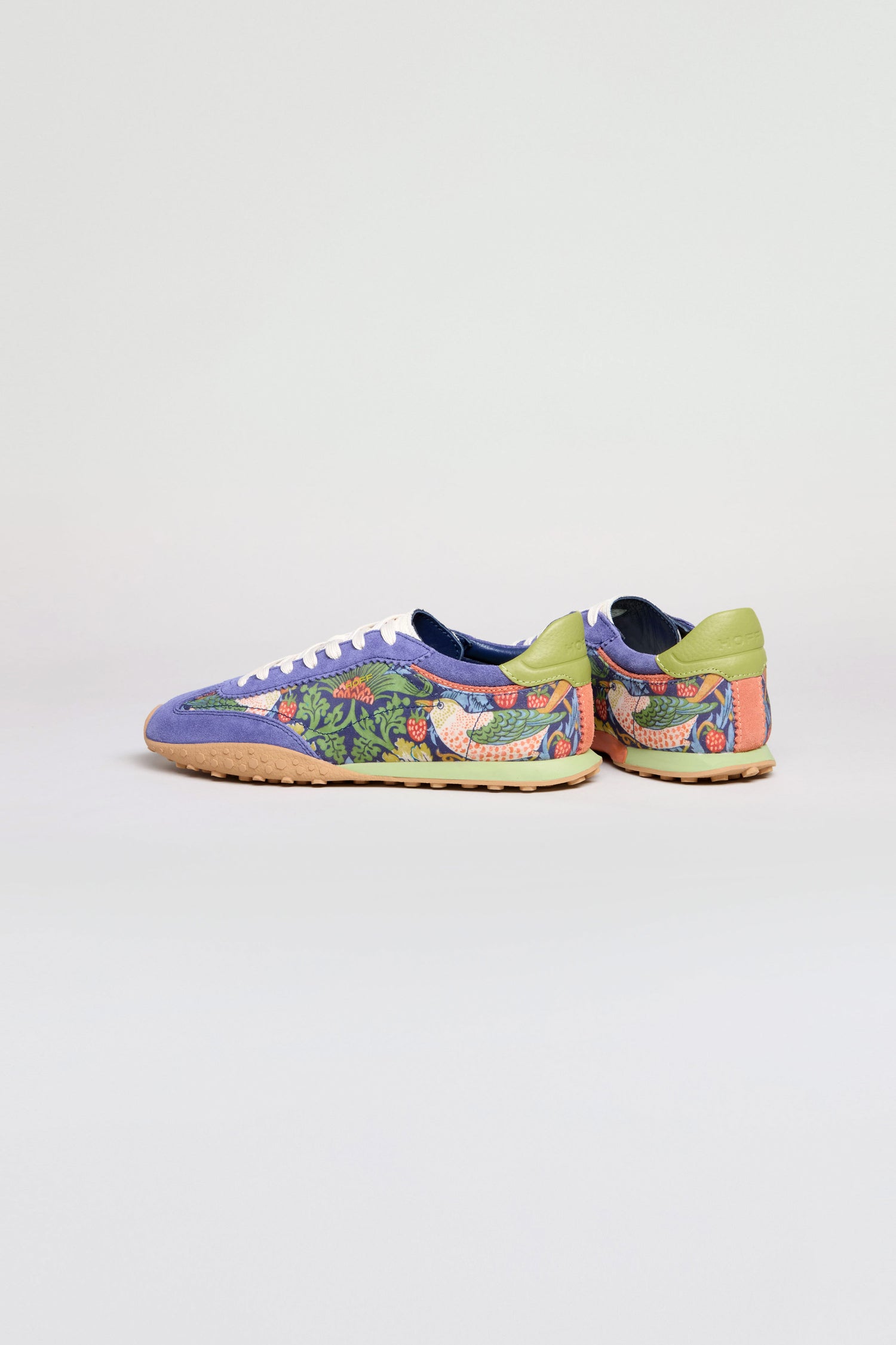 The William Morris Bridge Trainers are women’s leather shoes with a green, purple, and tan floral and bird print inspired by Strawberry Thief, featuring white laces, light brown soles, and shown from the back and side.