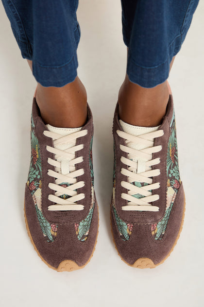 A person wears William Morris Bridge Trainers with Strawberry Thief print, beige laces, and blue pants, standing on a light surface.