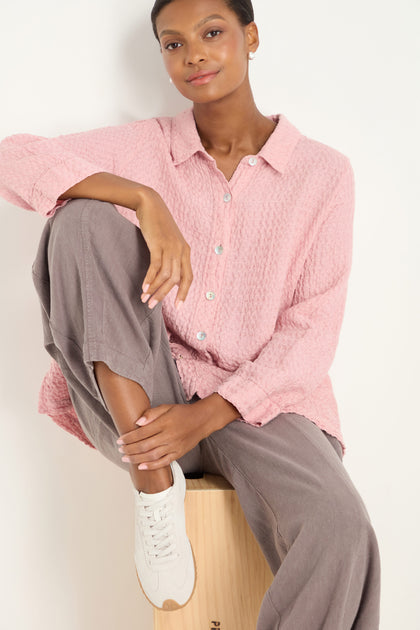 A person sits on a wooden box, wearing a textured pink button-up shirt, taupe pants, and white Bridge Suede Trainers, looking at the camera.