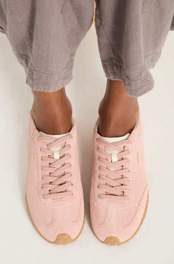 A person wearing loose gray pants and Bridge Suede Trainers—light pink with retro-inspired design and tan rubber soles—stands on a white surface.