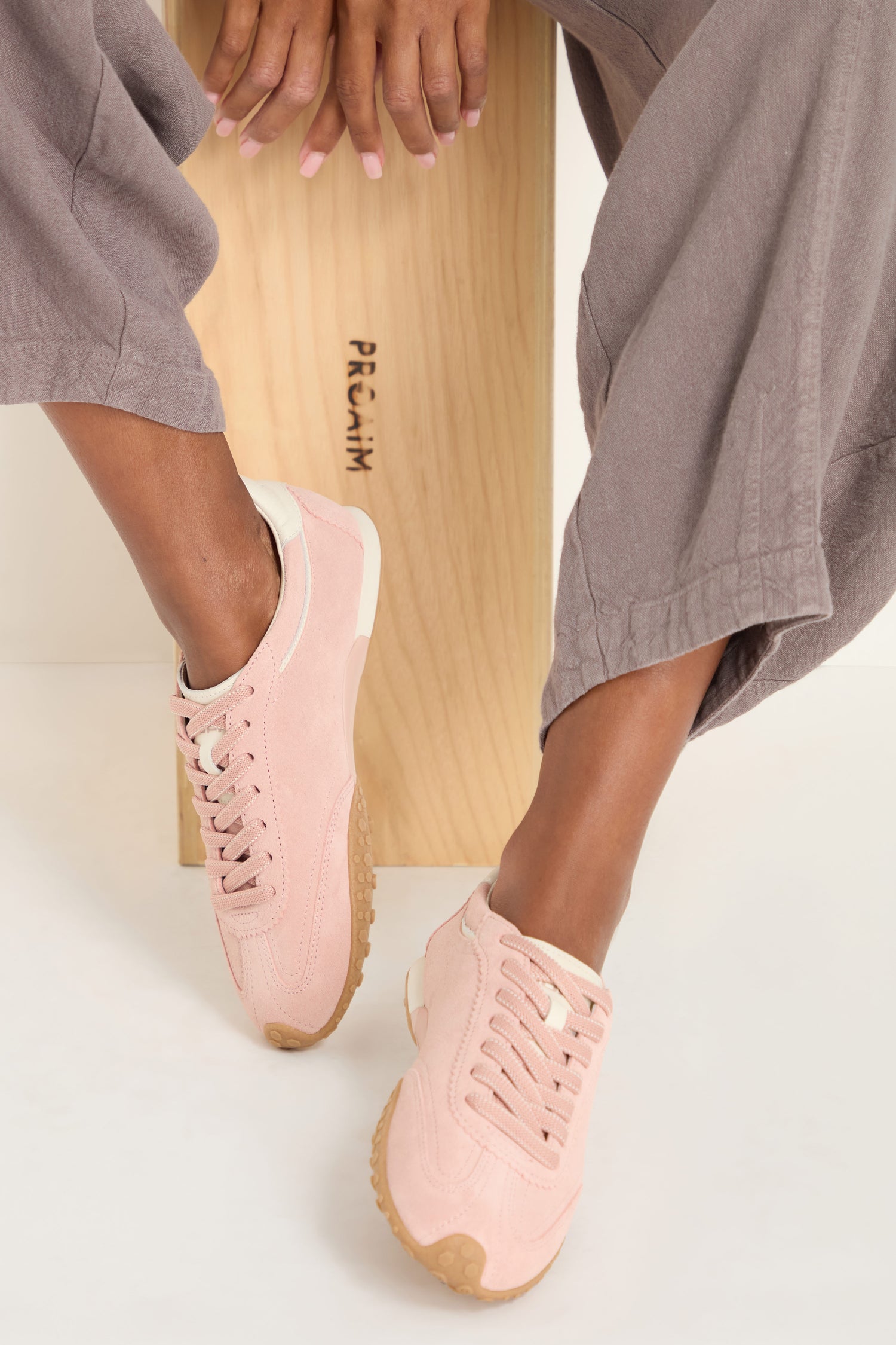 Person sitting on a wooden bench wearing mauve pants and Bridge Suede Trainers—light pink sneakers with beige soles—feet crossed at the ankle.