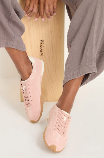 Person sitting on a wooden bench wearing mauve pants and Bridge Suede Trainers—light pink sneakers with beige soles—feet crossed at the ankle.