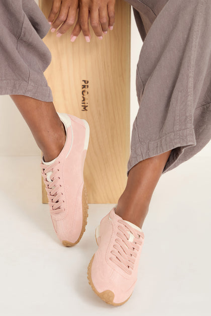Person sitting on a wooden bench wearing mauve pants and Bridge Suede Trainers—light pink sneakers with beige soles—feet crossed at the ankle.