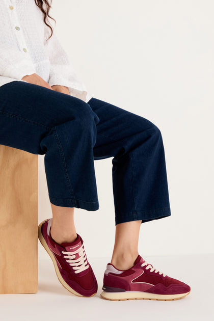 A person seated on a wooden block wears a white shirt, dark blue jeans, and City Trainers—premium suede footwear with white laces—against a plain light background.