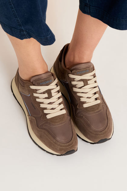 Person wearing City Trainers—premium suede shoes with cream-colored laces—paired with navy pants, standing on a light surface.