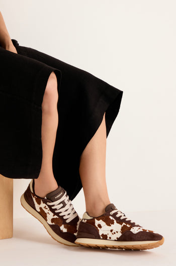A person wearing a long black skirt sits on a wooden block, showcasing the Animal Print City Trainers—premium suede sneakers featuring a brown and white cowhide pattern with beige soles—against a plain background.