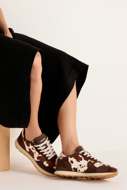 A person wearing a long black skirt sits on a wooden block, showcasing the Animal Print City Trainers—premium suede sneakers featuring a brown and white cowhide pattern with beige soles—against a plain background.