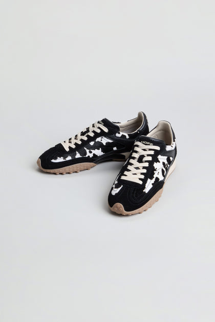 A pair of Animal Print Bridge Trainers featuring a black and white camo pattern in low-top suede, beige laces, and tan rubber soles, displayed on a plain light background.