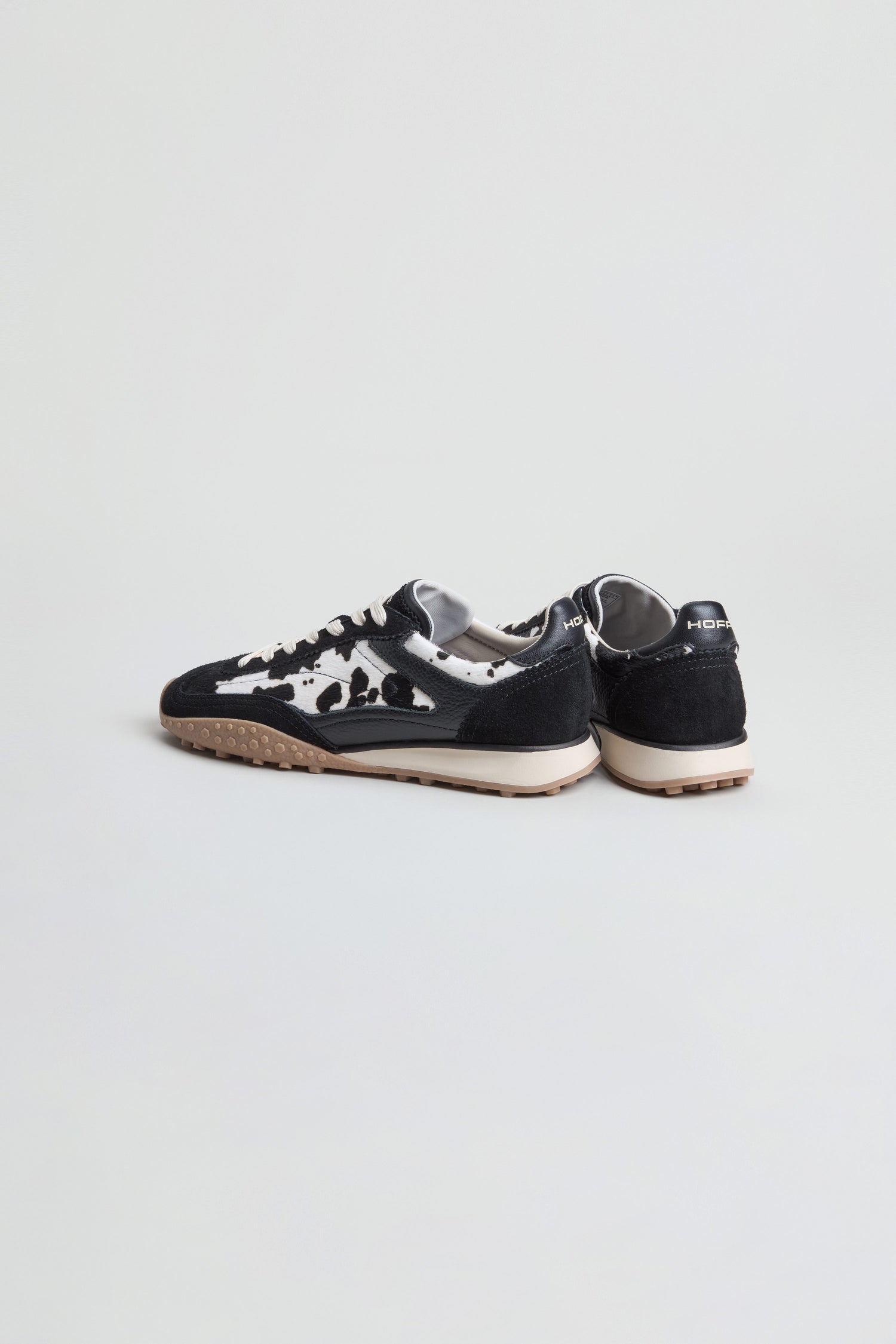 A pair of Animal Print Bridge Trainers featuring a bold black and white cow print, tan rubber soles, and black heel tabs displayed on a plain white background.