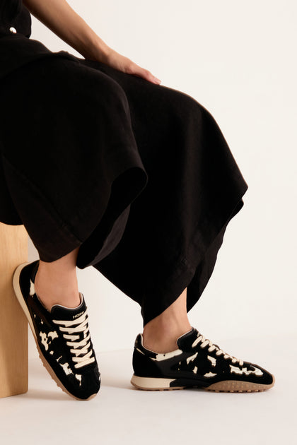 A person, seated on a wooden stool and dressed in a black skirt, showcases the Animal Print Bridge Trainers against a plain white background.