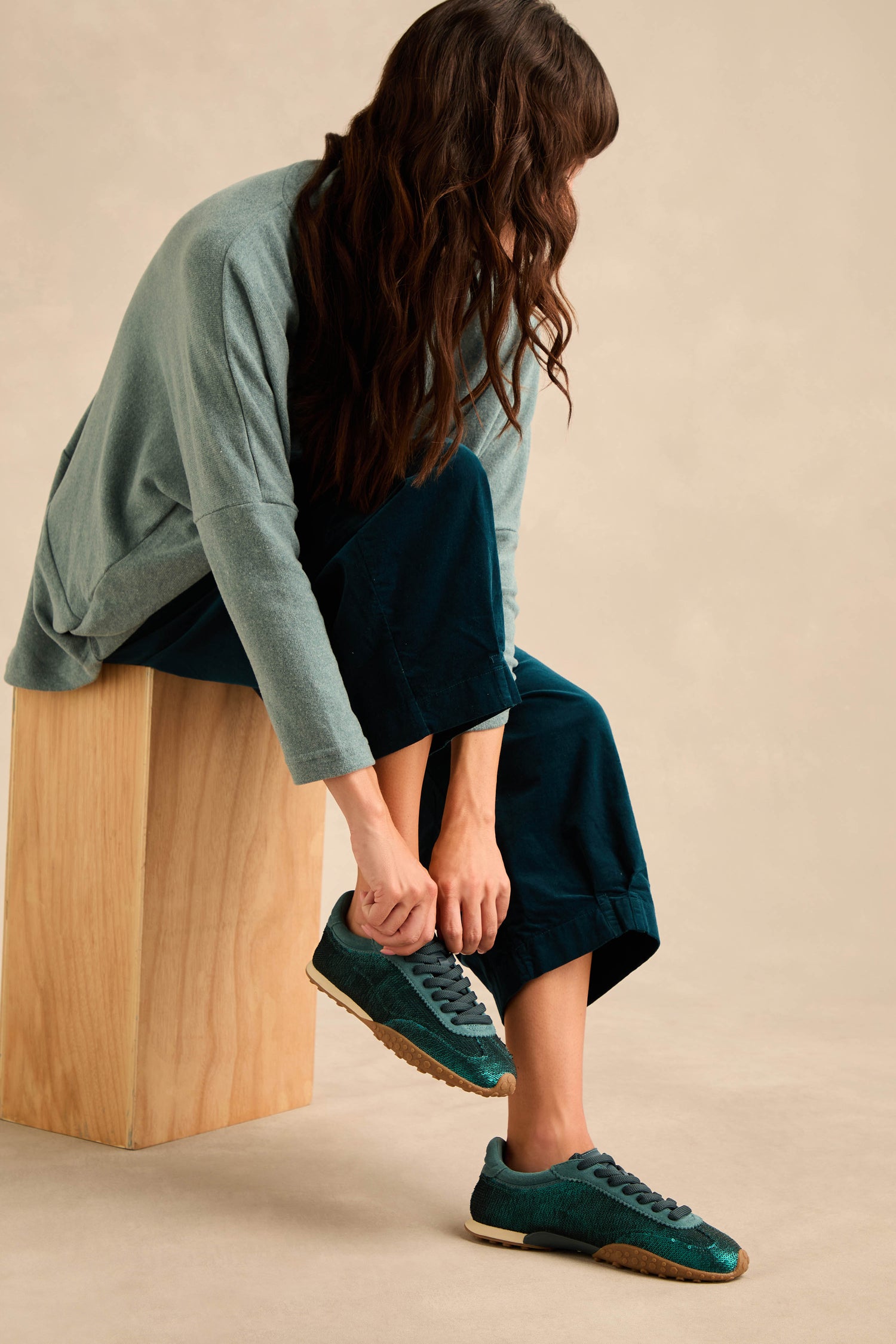 A person is sitting on a wooden block, wearing dark blue pants and a light teal top, tying Sequin Bridge Trainers against a neutral background.