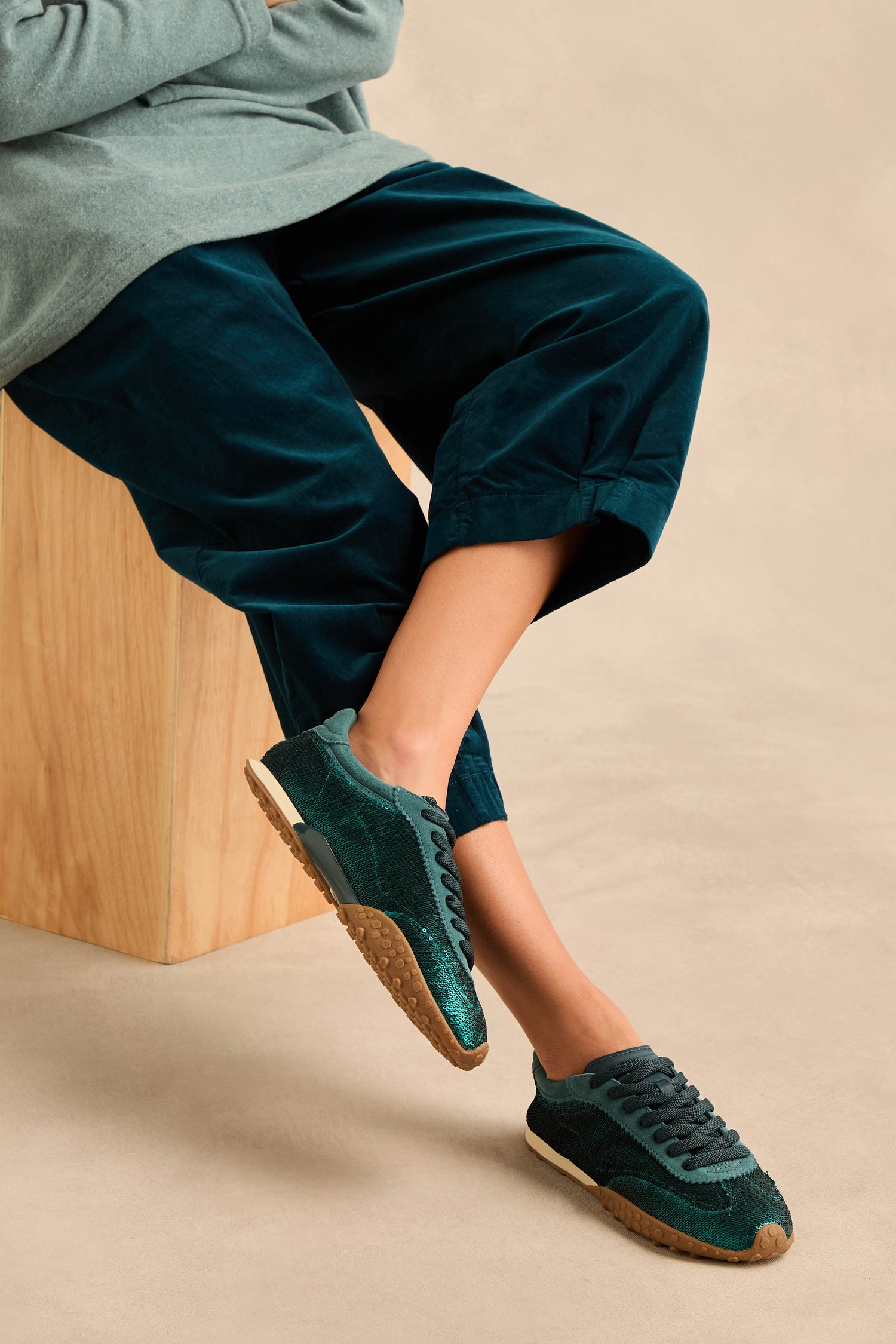 Person sitting on a wooden block, wearing dark green pants and Sequin Bridge Trainers—green sneakers with gum soles and a comfortable memory-foam insole—with their legs crossed.