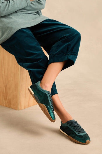 Person sitting on a wooden block, wearing dark green pants and Sequin Bridge Trainers—green sneakers with gum soles and a comfortable memory-foam insole—with their legs crossed.