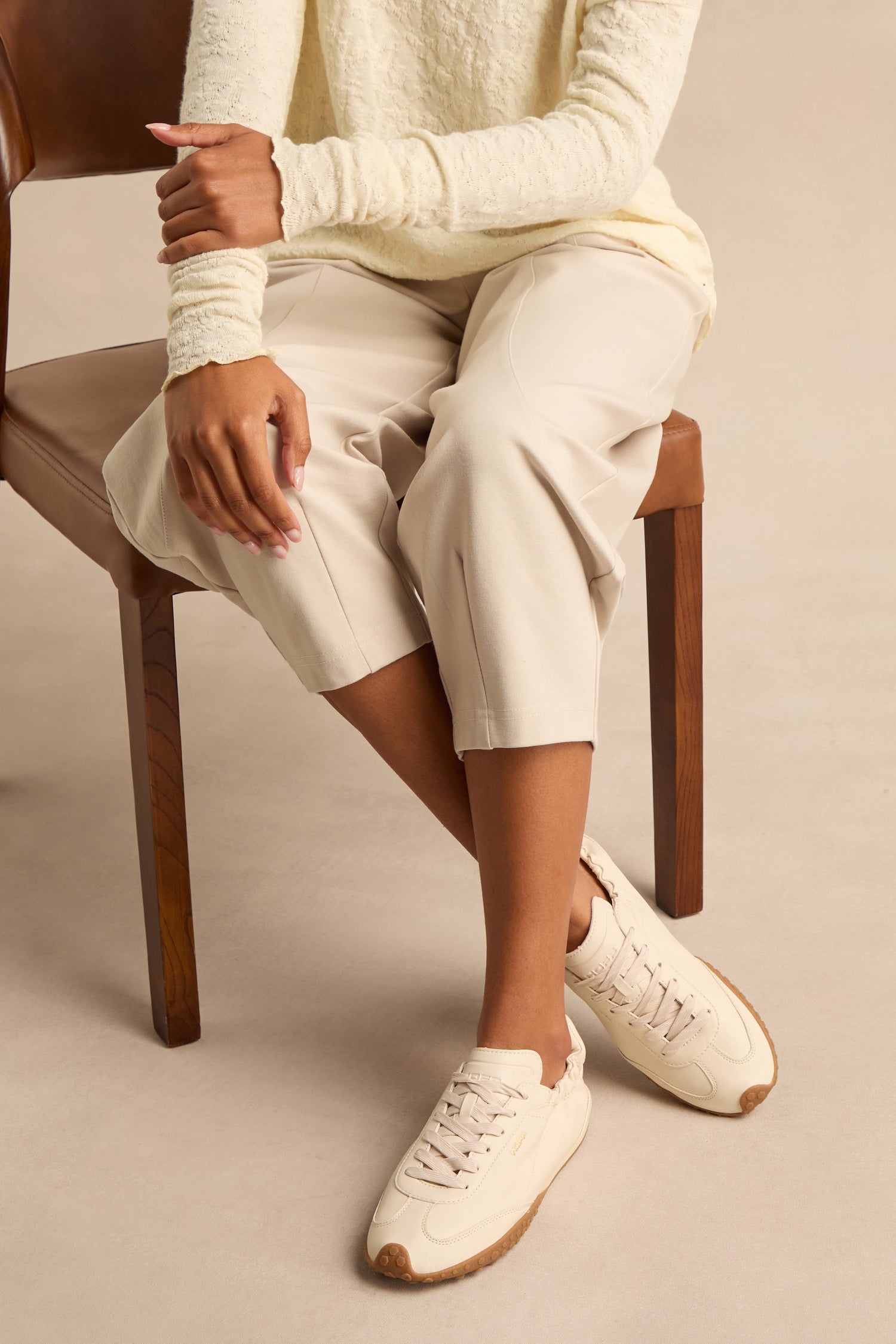 A person sits on a wooden chair, dressed in a cream long-sleeve top and light beige pants, wearing Napa Leather Bridge Trainers—low profile cream leather shoes with tan soles. Their hands rest on one knee.