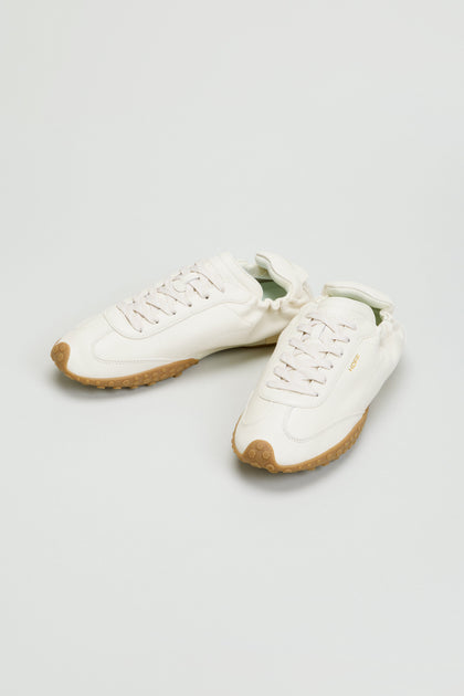 A pair of Napa Leather Bridge Trainers in white, featuring low-top design, lace-up closures, and gum rubber outsoles, displayed against a plain light gray background.