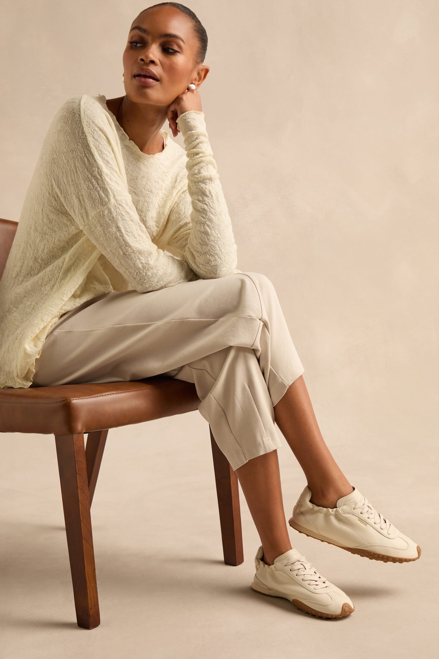 A person sits on a brown chair, wearing a textured cream sweater, light beige pants, and elegant Napa Leather Bridge Trainers in cream against a neutral background.
