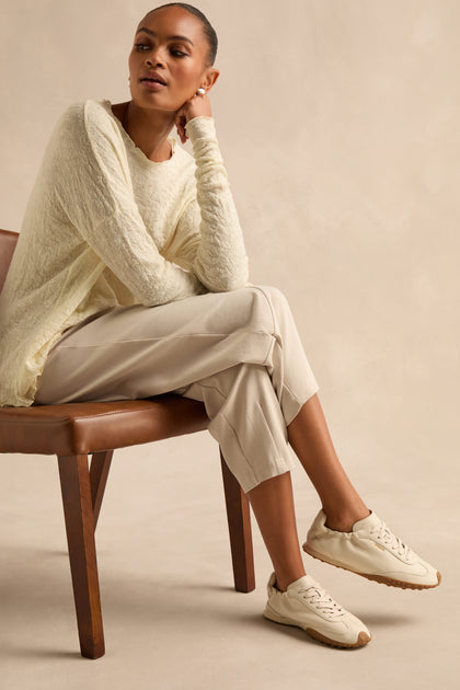 A person sits on a brown chair, wearing a textured cream sweater, light beige pants, and elegant Napa Leather Bridge Trainers in cream against a neutral background.