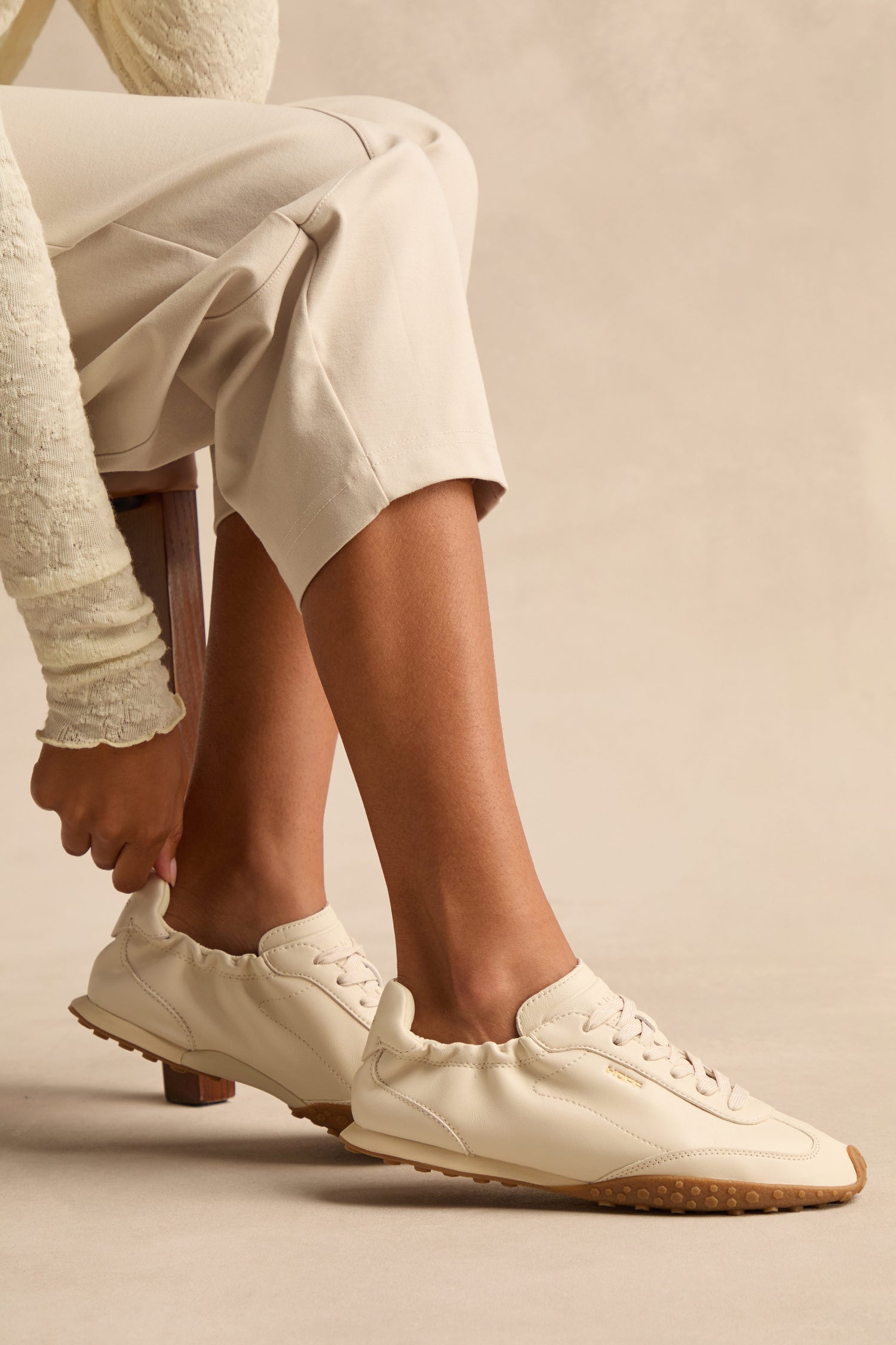 A person sits on a bench wearing light-colored pants and cream Napa Leather Bridge Trainers with gum soles, reaching to adjust the heel of one shoe.