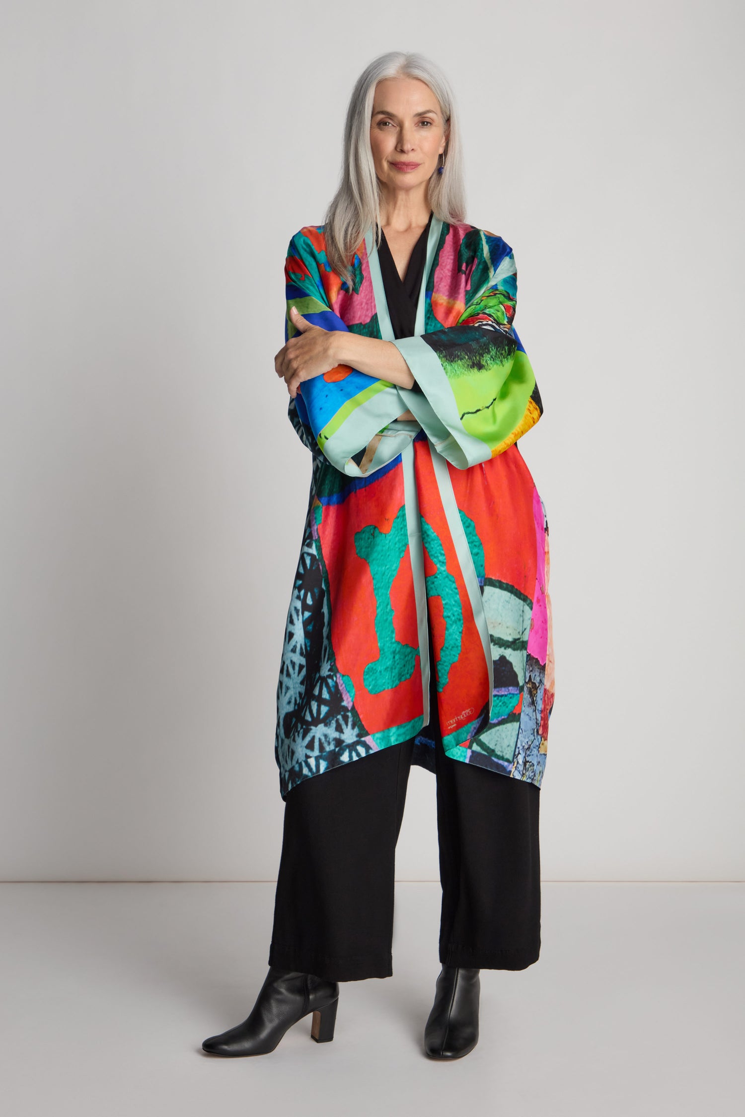 A woman with long white hair stands in front of a plain background, wearing a colorful, oversized Ibiza Silk Kimono by Isabelle Gougenheim, along with a black top, black pants, and black heeled boots.