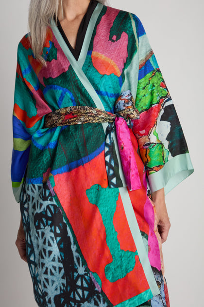 A person wearing a vibrant Ibiza Silk Kimono, adorned with abstract patterns and secured with a belt, standing against a plain background.