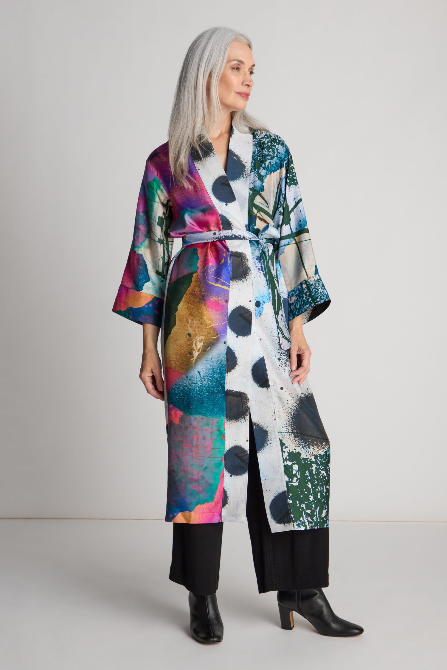 A woman with long gray hair stands wearing the Connect the Dots Long Silk Kimono by Isabelle Gougenheim over black pants and black ankle boots. She is facing slightly to the side with a neutral expression.