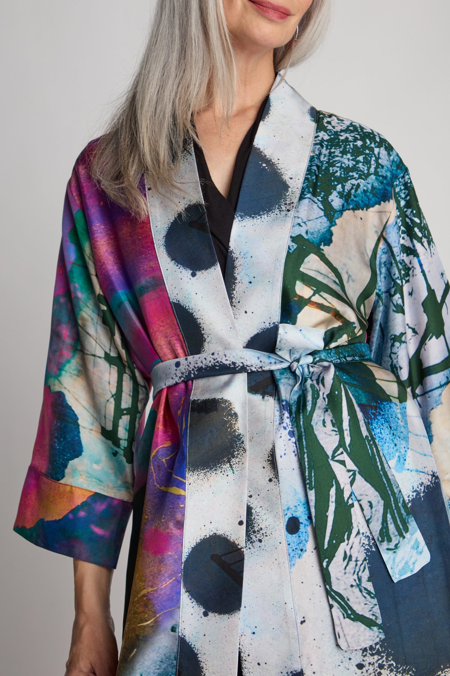 A woman with long, gray hair is wearing a Connect the Dots Long Silk Kimono tied at the waist, showcasing its colorful, patterned design. Only her upper torso is visible.