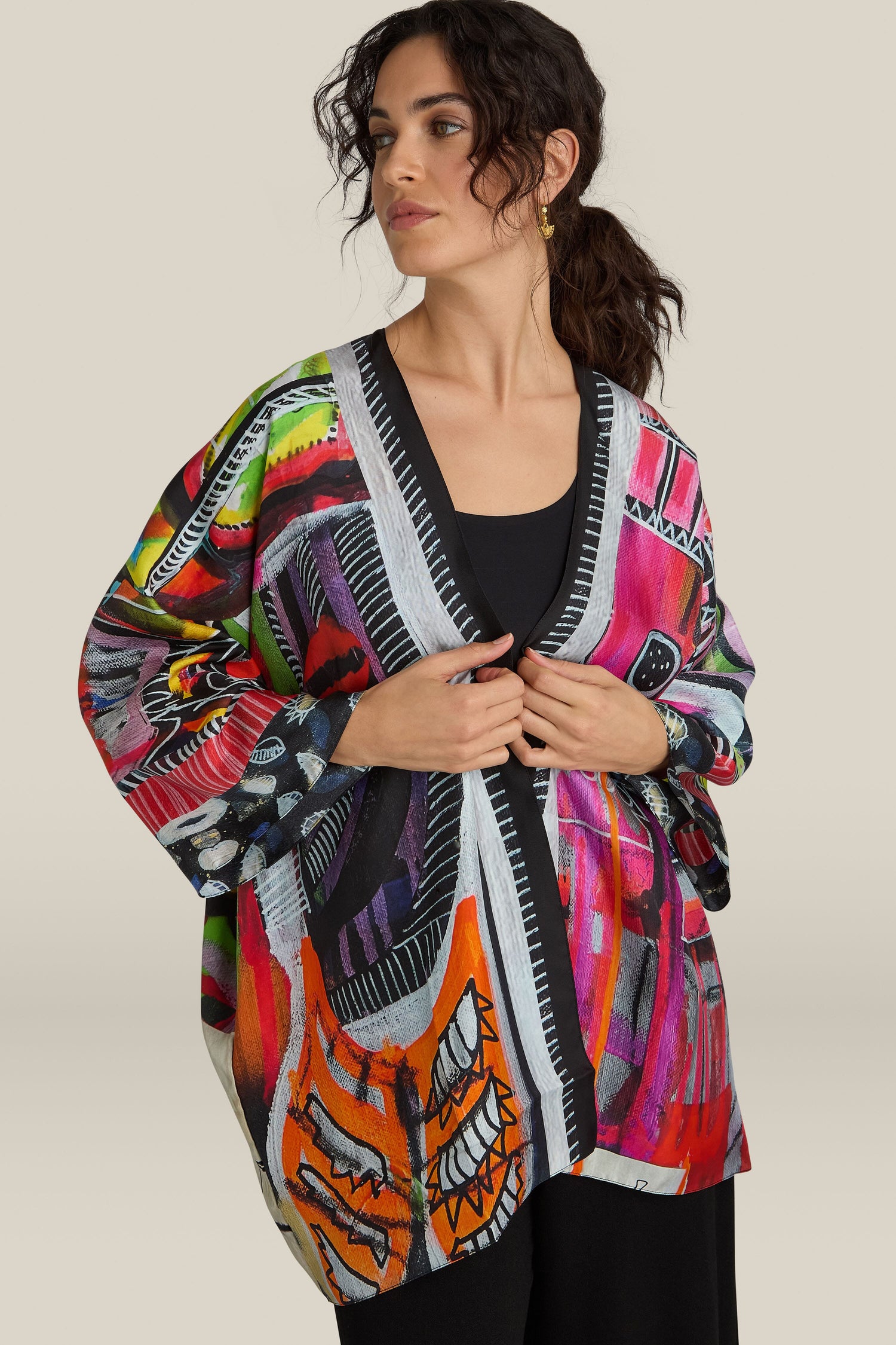 A person stands wearing a vibrant Havana Print Short Silk Kimono over a black top, with dark hair pulled back and both hands adjusting the garment.