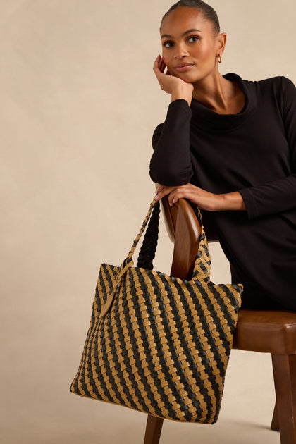 A woman in a black dress sits on a chair, resting her elbow on the backrest, holding the Reversible Braided Shopper—a spacious handbag featuring a striking black and tan checker pattern.