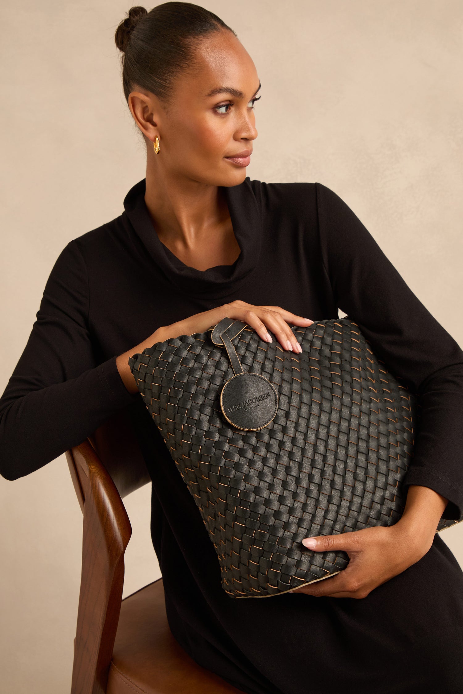 Woman in a black dress sits on a wooden chair, holding a large woven reversible bag with a round tag.