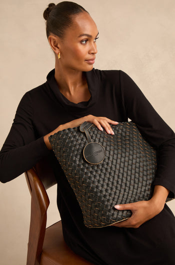 Woman in a black dress sits on a wooden chair, holding a large woven reversible bag with a round tag.