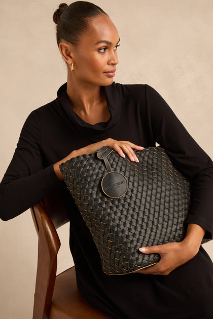 Woman in a black dress sits on a wooden chair, holding a large woven reversible bag with a round tag.
