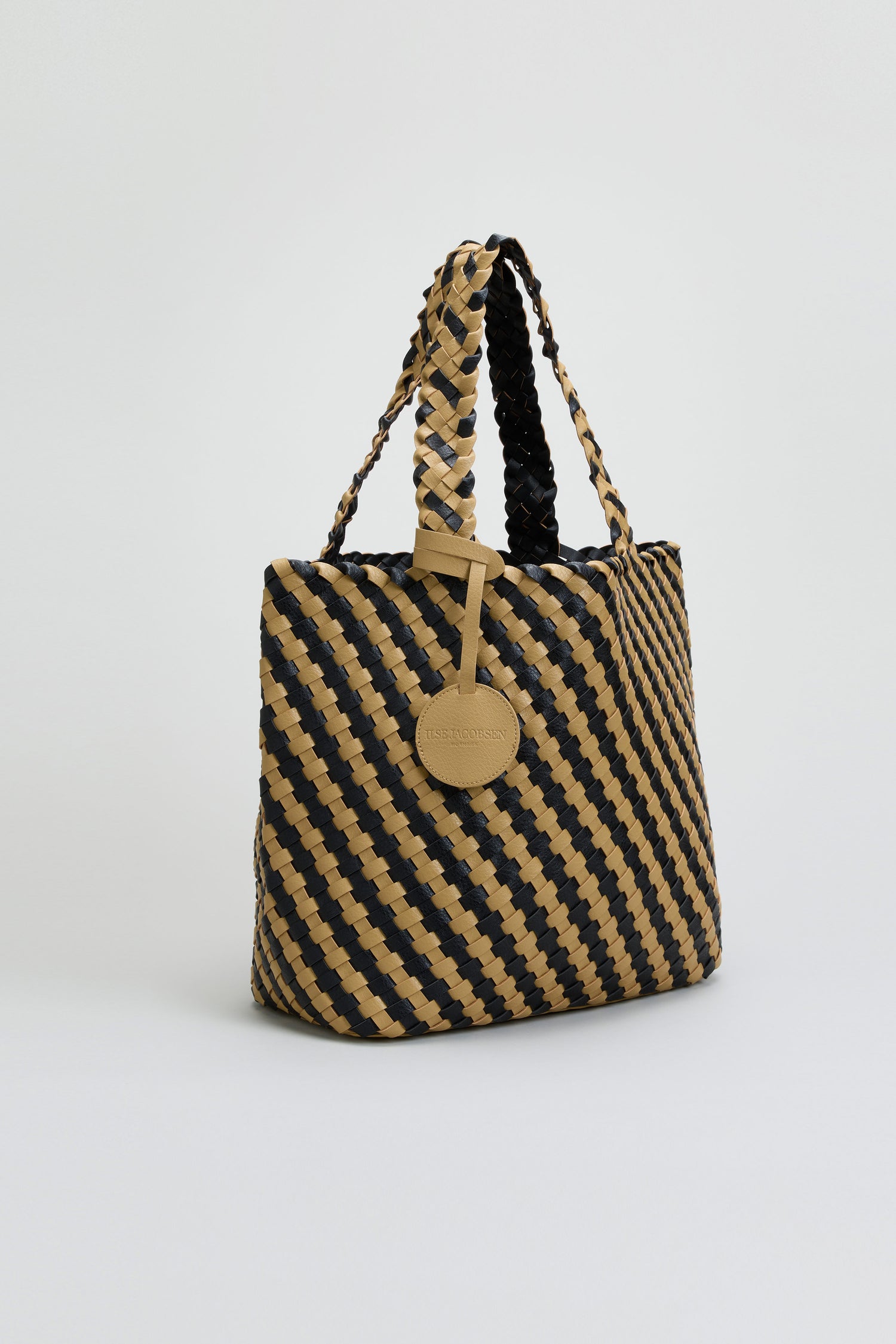 The Reversible Braided Shopper showcases a diagonal black and tan basket weave pattern, braided handles, and a round beige tag hanging from one handle, photographed against a plain background.