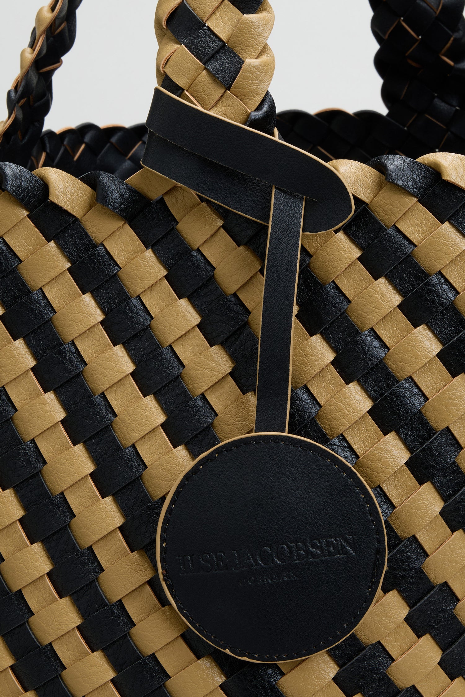 Close-up of the Reversible Braided Shopper, featuring a woven black and tan basket weave design and a round black leather tag labeled “ILSE JACOBSEN HORNBÆK.”.