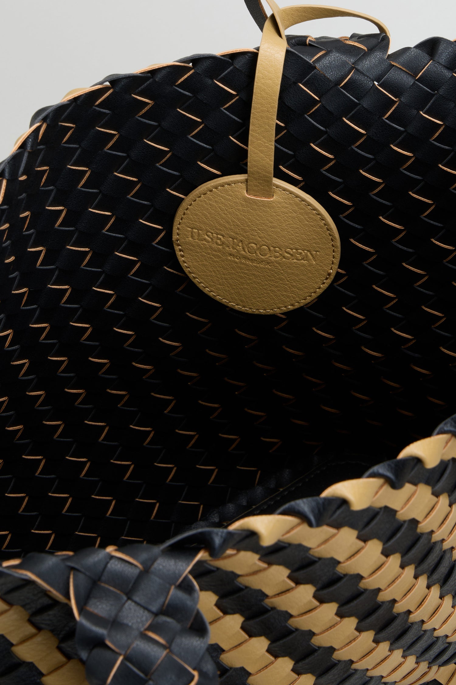 Close-up of a basket weave handbag in black and tan, featuring a round tan leather tag embossed with “ILSE JACOBSEN.”.
