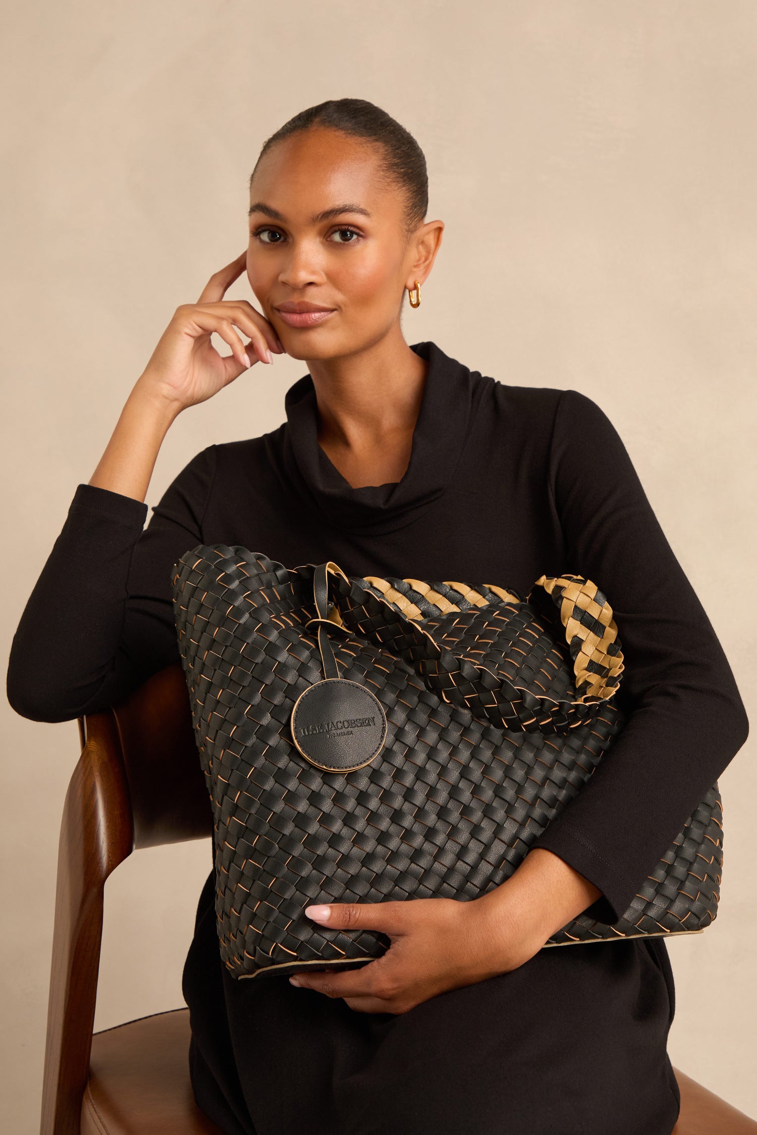 A woman in a black dress sits on a wooden chair, holding the Reversible Braided Shopper with a circular tag, looking at the camera.