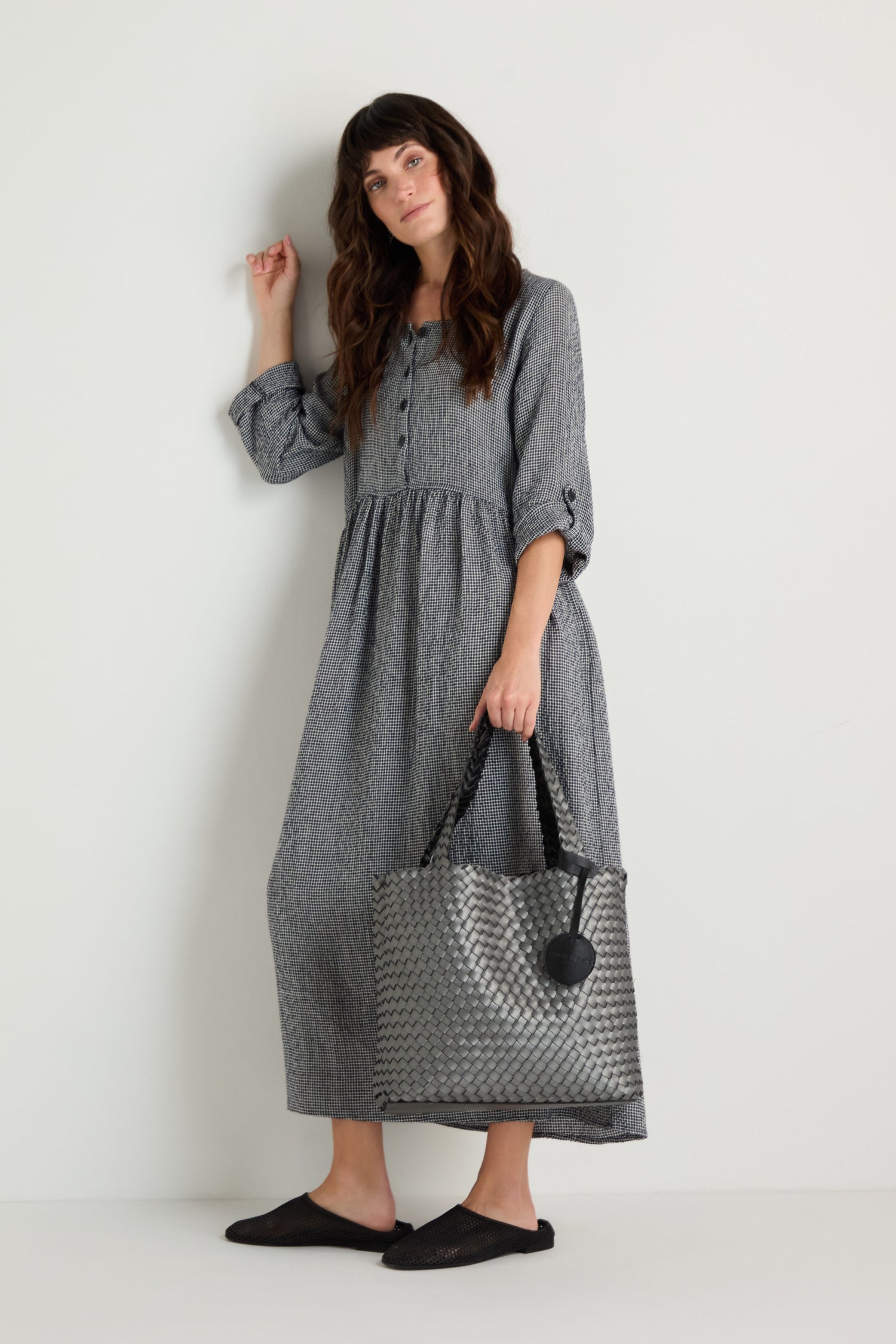 A woman in a long-sleeved, button-up gray dress stands against a white wall, holding the Woven Reversible Bag and wearing black slip-on shoes.