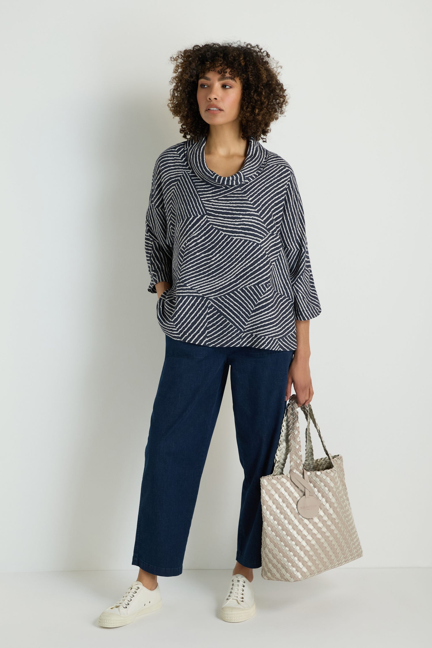 A woman stands in front of a plain wall, wearing a striped sweater, wide-leg jeans, white sneakers, and holding a Woven Reversible Grey & Silver Bag.