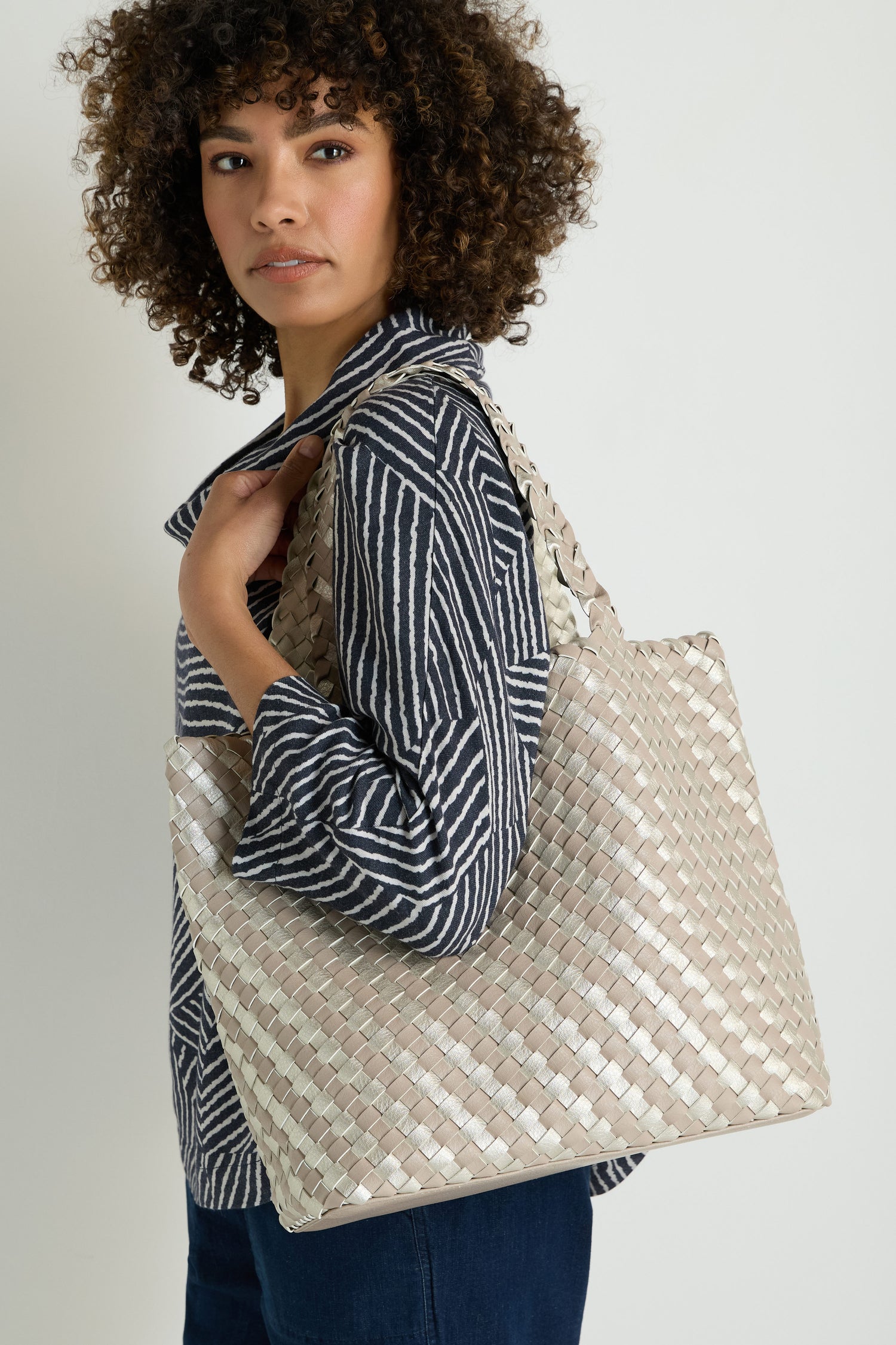 Woman with curly hair in a striped shirt and dark pants carries the Woven Reversible Grey & Silver Bag over her shoulder.