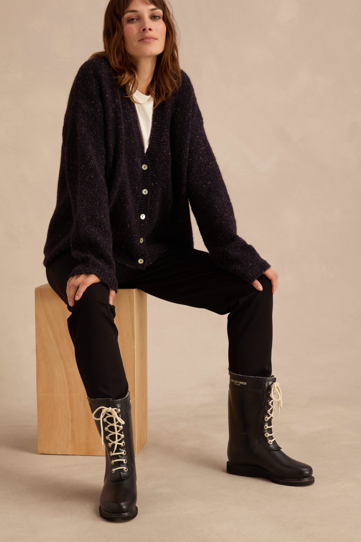 A woman sits on a wooden block against a beige background, dressed in a dark cardigan and black pants, showcasing the Lace Up Wellington Boots—ideal wet-weather footwear.